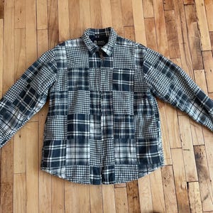 May include: A long-sleeved shirt with a button-down front and a patchwork design. The shirt features a mix of plaid patterns in navy, white, and grey. The shirt is laid flat on a wooden surface.