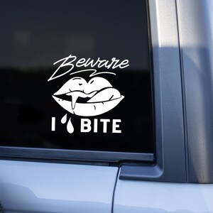 May include: White vinyl sticker with a black background. The sticker features a stylized image of a mouth with fangs and a tongue sticking out. The text "Beware I Bite" is below the image.