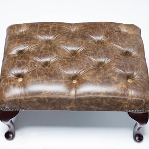 May include: A brown leather tufted ottoman with four wooden legs. The ottoman has a distressed look and is upholstered in a brown leather.