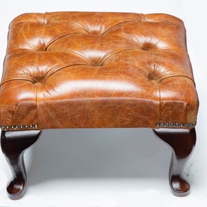 May include: A brown leather ottoman with a tufted top and four wooden legs. The ottoman has a distressed look and is perfect for adding a touch of rustic charm to any room.