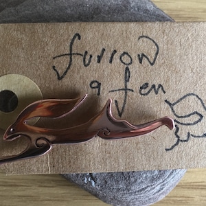 May include: A copper-coloured metal hare pin on a brown tag with the handwritten text "furrow & fen". The pin is a stylized depiction of a running hare. The tag has a circular hole and is resting on a grey stone.