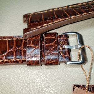 May include: A brown leather watch strap with a crocodile pattern and a silver buckle. The strap has a stitched edge and is folded over on itself.