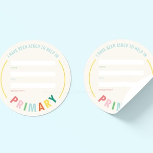 May include: Two round stickers with the text "I HAVE BEEN ASKED TO HELP IN" at the top. The stickers have spaces for "name", "date", and "assignment". The word "PRIMARY" is at the bottom in colorful letters.