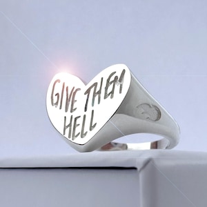 Op de afbeelding: Een zilveren ring in de vorm van een hart met de woorden "GIVE THEM HELL" gegraveerd op de voorkant.