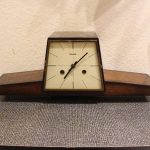 Può includere: Un orologio vintage in legno con quadrante color crema e lancette nere. L'orologio ha una forma trapezoidale con finitura marrone scuro ed è posto su una base rettangolare con gambe color ottone. Sul quadrante dell'orologio è stampata la parola "Hama".