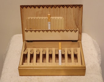 Puede incluir: Una caja de madera para cigarrillos con tapa abatible y diseño decorativo. La caja tiene 10 ranuras para cigarrillos y un compartimento separado para un encendedor o cerillas.