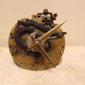 Könnte beinhalten: Nahaufnahme eines Uhrwerks mit goldfarbenen Zeigern und Zahnrädern. Das Zifferblatt der Uhr ist aus Messing und hat ein abgenutztes, gealtertes Aussehen.