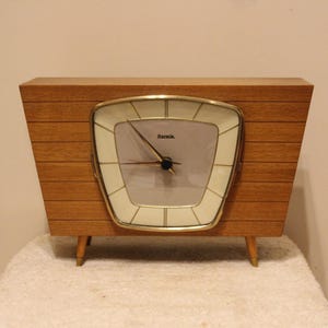 Op de afbeelding: Een vintage houten klok met een witte wijzerplaat en gouden afwerking. De klok heeft een unieke, licht gebogen vorm en heeft het woord "Hamile" op de wijzerplaat.
