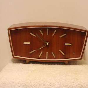 May include: A vintage wooden clock with a brown finish and a cream-colored border. The clock face features gold-colored markers and hands. The clock is rectangular with a curved top and sits on small wooden legs.