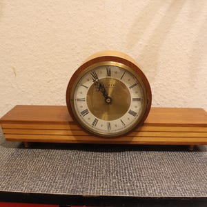 May include: A wooden mantel clock with a round, white face and black Roman numeral hour markers. The clock has a gold-coloured trim and hands.