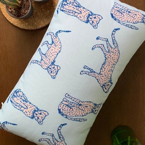 May include: A light blue throw pillow with a repeating pattern of pink cheetahs printed on it. The cheetahs are outlined in blue.