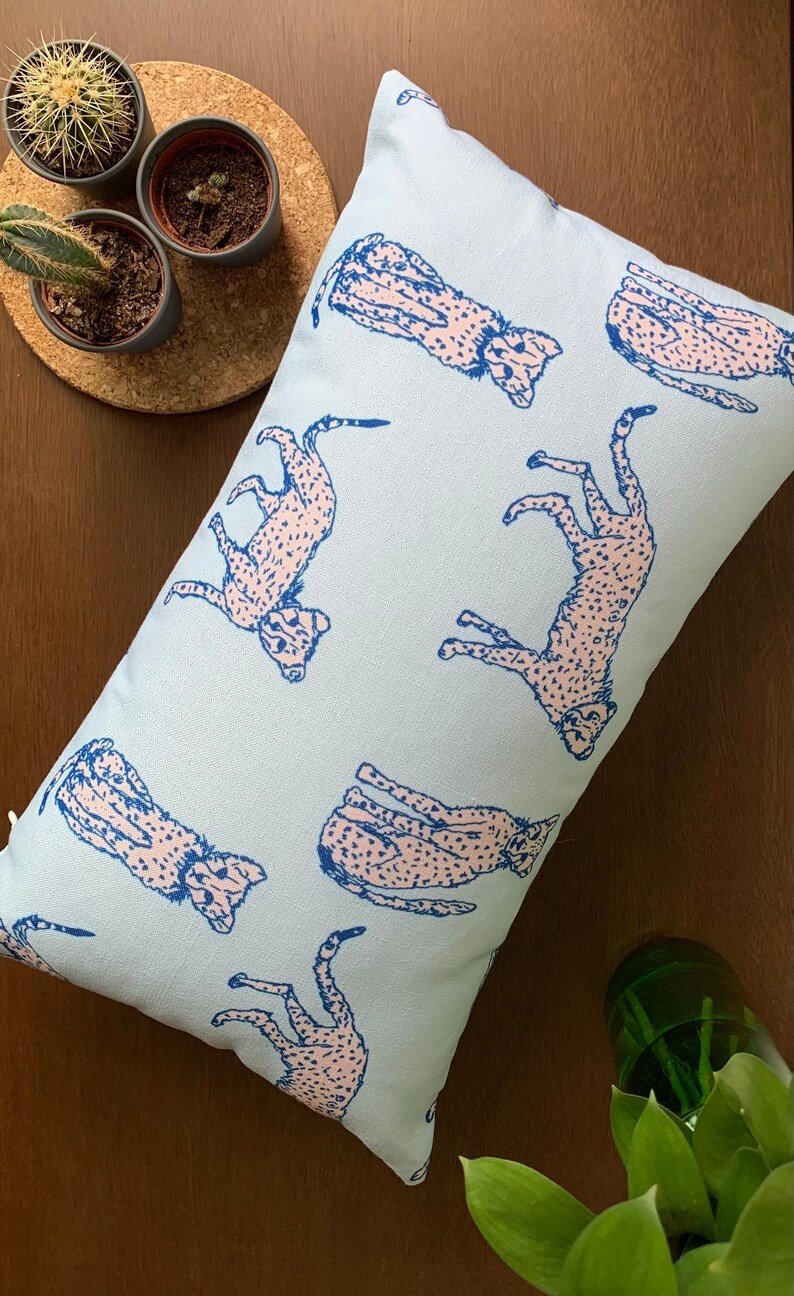 May include: A light blue throw pillow with a repeating pattern of pink cheetahs printed on it. The cheetahs are outlined in blue.