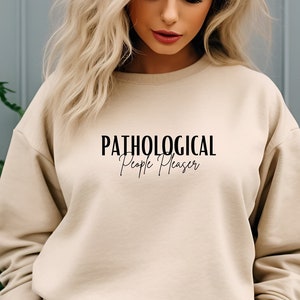 Könnte beinhalten: Ein beiger Pullover mit dem Aufdruck "PATHOLOGICAL People Pleaser" auf der Vorderseite.