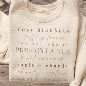 May include: Cream-colored sweatshirt with the words "cozy blankets, falling leaves, football cheers, pumpkin lattes, knit scarves, apple orchards, chunky sweaters, happy hearts" printed on the front. A cup of coffee and a donut are on the left.