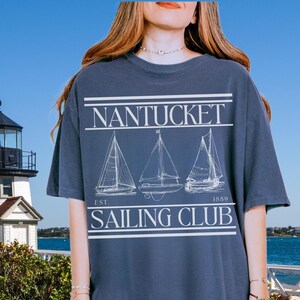 Peut inclure: T-shirt bleu marine avec l'inscription blanche "Nantucket Sailing Club". Le design comprend trois illustrations de voiliers et l'année "1889". Le t-shirt est de coupe décontractée.