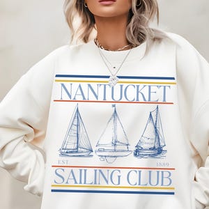 Peut inclure: Sweat-shirt crème avec l'inscription "NANTUCKET SAILING CLUB" et trois illustrations de voiliers. Le texte est au-dessus et en dessous des voiliers, avec l'année "1889" incluse. Le design présente des rayures bleues, jaunes et rouges.