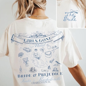 Puede incluir: Camiseta color crema con un diseño gráfico azul. El diseño incluye el texto "Girls Gone Mild" y "Bride & Prejudice" con ilustraciones de una novia, regalos y otros elementos temáticos de boda. El texto "The Bride" está en la parte superior de la espalda.