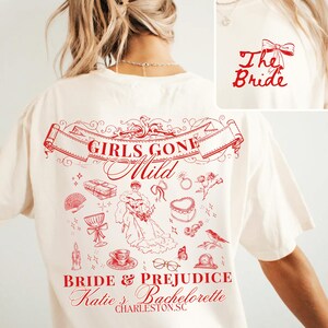Op de afbeelding: Crèmekleurig T-shirt met rode afbeelding. De achterkant heeft de tekst "GIRLS GONE Mild" in een banner, met illustraties van een bruid, cadeaus en andere elementen met een bruiloftthema. De tekst "BRIDE & PREJUDICE Katie's Bachelorette CHARLESTON, SC" is ook zichtbaar.