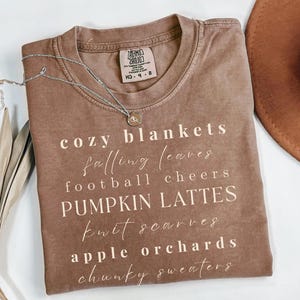 Op de afbeelding: Een bruin t-shirt met de woorden "cozy blankets, falling leaves, football cheers, pumpkin lattes, knit scarves, apple orchards, chunky sweaters" in het wit gedrukt. Een zilveren ketting en een bruine hoed zijn ook zichtbaar.