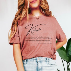 May include: A dusty rose-colored t-shirt with the word "Kahu" in a stylish script, accompanied by a definition. The definition describes a guardian, keeper of animals, and protector. The text also includes the pronunciation and a reference to animal lovers.