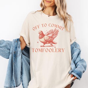 May include: A cream-colored t-shirt with the text "Off to commit tomfoolery" in red lettering. A stylized illustration of a dove with its wings spread is centered above the text.