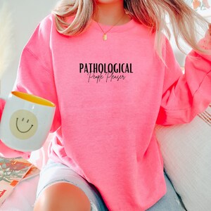 May include: A pink sweatshirt with the text "Pathological People Pleaser" in black.
