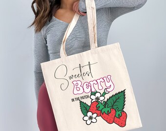 Puede incluir: Una bolsa de tela blanca con el texto "Sweetest Berry in the Patch" y un gráfico de fresas y flores.