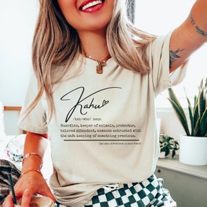 May include: Cream-colored t-shirt with the word "Kahu" in a cursive font, along with a definition. The definition describes a guardian, keeper of animals, and protector. The shirt also includes the phrase "See also: animal lover or best friend."