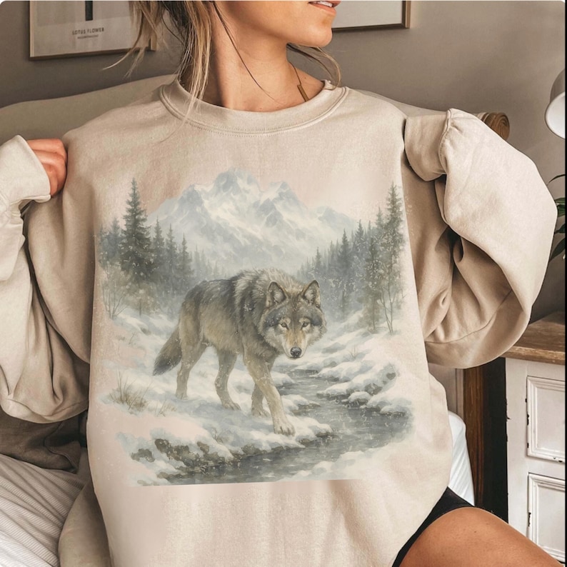 Puede incluir: Sudadera beige con una ilustraci&oacute;n detallada de un lobo caminando por un paisaje nevado. El lobo es gris y marr&oacute;n, con &aacute;rboles y monta&ntilde;as en el fondo, evocando un tema invernal.