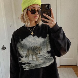 Puede incluir: Sudadera negra con un gr&aacute;fico detallado de un lobo caminando en un bosque nevado con monta&ntilde;as al fondo. La sudadera tiene cuello redondo y mangas largas. La persona de la imagen lleva un gorro verde ne&oacute;n y gafas de sol.