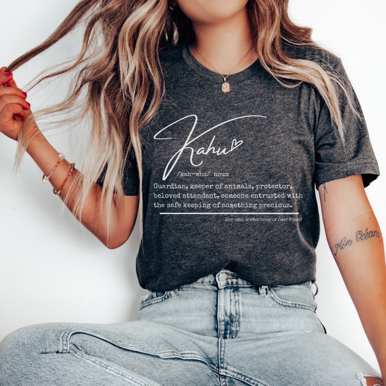 May include: Dark gray t-shirt with the word "Kahu" in a cursive font. Below the word is a definition: "Guardian, keeper of animals, protector, beloved attendant, someone entrusted with the safe keeping of something precious."