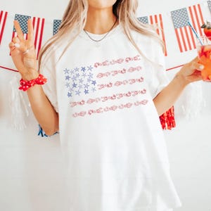 May include: White t-shirt with a red, white, and blue American flag design. The flag is made up of blue stars and red and white seashells.