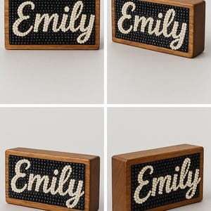 May include: Wooden block sign with the name "Emily" spelled out in cream-colored beads on a black beaded background. The sign is encased in a rectangular, brown wooden frame.