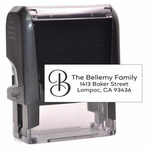 May include: A black self-inking stamp with a white label. The label features the text "The Bellemy Family" and an address, along with a decorative initial "B". The stamp is designed for repeated use.