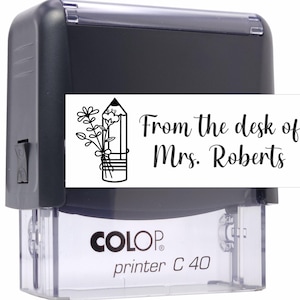 May include: Black and white stamp with a pencil and flowers design. The text reads "From the desk of Mrs. Roberts". The stamp is labeled "COLOP printer C 40".