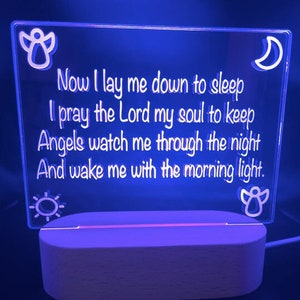 May include: A white LED night light with a wooden base. The light features a clear acrylic panel with the words "Now I lay me down to sleep I pray the Lord my soul to keep Angels watch me through the night And wake me with the morning light." The panel also features a crescent moon, a sun, and two angels.