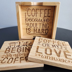May include: Set of four wooden coasters with engraved sayings about coffee. The coasters are in a holder with the saying "Coffee because adulting is hard".