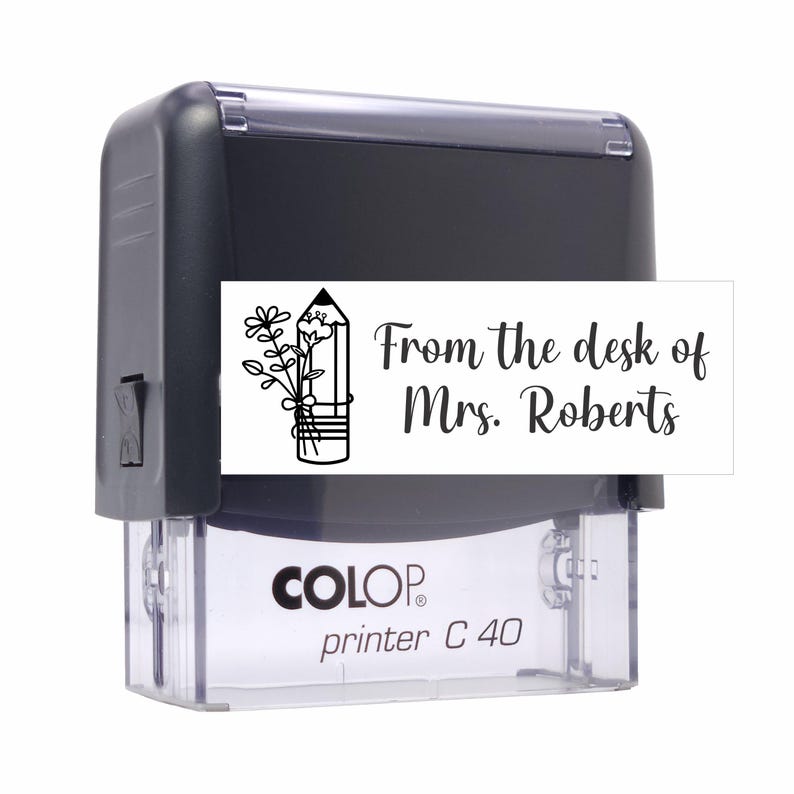 May include: A black self-inking rubber stamp with a clear base labeled Colop printer C 40. The stamp features a white rectangular label with a black line drawing of a pencil with flowers and the text From the desk of Mrs. Roberts.