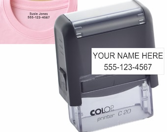 May include: A black self-inking stamp labeled Colop Printer C 20 with a clear base. The stamp displays the text YOUR NAME HERE 555-123-4567. A pink t-shirt collar is visible in the background with the text Susie Jones 555-123-4567 printed on it.