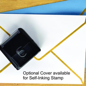 May include: A black self-inking stamp positioned on a white envelope with gold geometric lines, which is placed over a light blue envelope. The text on the white envelope reads: Optional Cover available for Self-Inking Stamp.