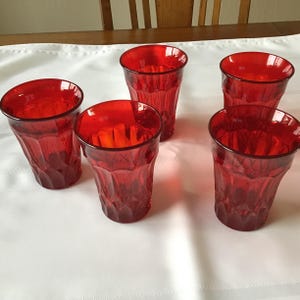May include: Five red drinking glasses with a textured, faceted design. The glasses are arranged on a white tablecloth, with a wooden table and chairs visible in the background. The glasses are empty and appear to be made of glass.