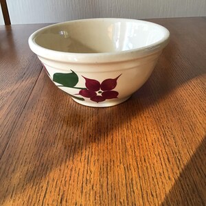 Puede incluir: Cuenco de cerámica color crema con un diseño floral. El cuenco presenta una flor granate con hojas verdes pintadas en el lateral. El cuenco está sobre una superficie de madera.