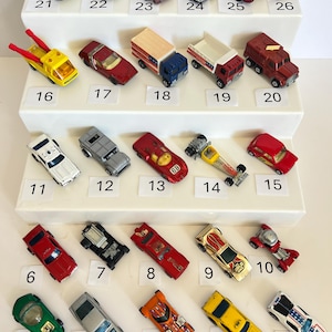 May include: A collection of twenty-six miniature die-cast toy cars arranged on a white tiered display stand. Each car is numbered from 1 to 26 with a small white label. The vehicles feature various colors, designs, and styles, including race cars, trucks, and sedans, all displayed in a grid-like formation.