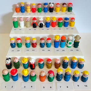 May include: A collection of fifty vintage plastic toy figures arranged on a four-tiered white display stand. Each figure features a simple cylindrical body with a painted face and is numbered from 1 to 50 on small white labels placed beneath them.