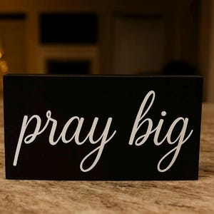 May include: A black rectangular wooden sign with the words "pray big" in white cursive lettering. The sign is placed on a textured surface, with a blurred background.