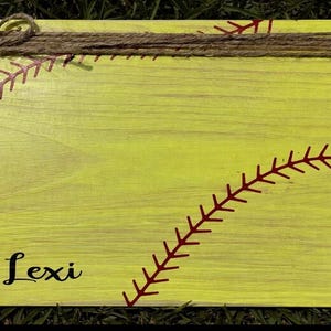 May include: A rectangular yellow wooden sign featuring red softball stitching patterns, a brown twine bow at the top, and black text that reads "Lexi" in the bottom left corner and "LADY CARDS" vertically along the right side of the board.