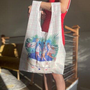 May include: A white reusable tote bag with a pink grid pattern. The bag features a colourful illustration of five people running through a tropical scene with palm trees and the words "Play" and "Pause" on a blue background.