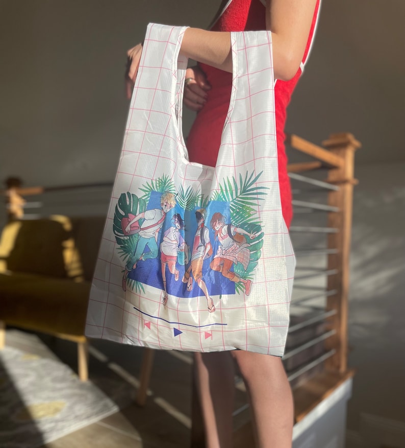 May include: A white reusable tote bag with a pink grid pattern. The bag features a colourful illustration of five people running through a tropical scene with palm trees and the words "Play" and "Pause" on a blue background.