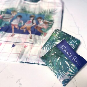 May include: A white and pink reusable shopping bag with a tropical print featuring four cartoon figures. The bag is folded and placed on a white marble surface. The bag is labelled "Fictional Skin".