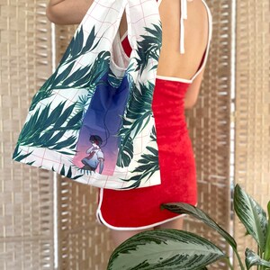 May include: A white reusable tote bag with a grid pattern and a tropical leaf print. The bag features a graphic of a person with headphones on, standing in front of a blue and purple background.